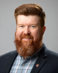 Headshot of Dr. Brian Luck. Dr. Luck is wearing a blue plaid collared shirt and a black coat.  He has medium length red hair and a full red beard.