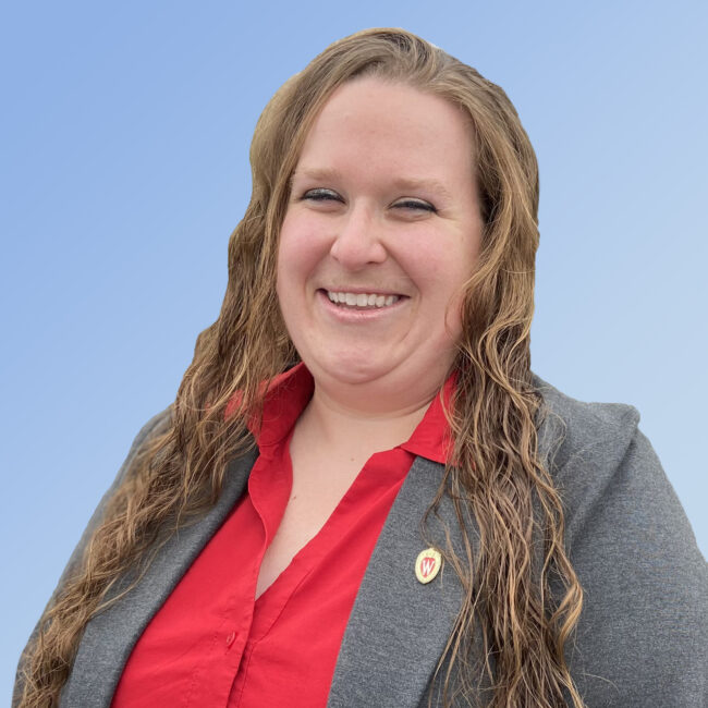 Photo of Lyssa Seefeldt.  Lyssa is wearing a red collared shirt and a gray blazer with a UW-Madison crest pin in the lapel.  She is wearing her long brown hair down and is standing in front of a sky blue background.